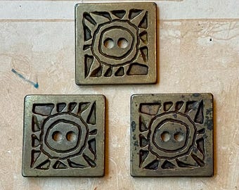May include: Three square, antique brass-colored buttons. Each button features a sun-like design with a central circle containing two holes for sewing. The buttons have a weathered, vintage appearance, suggesting they are old or have been aged.