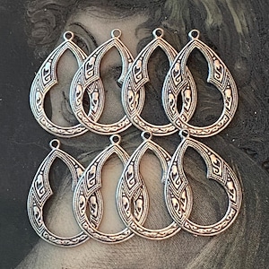 May include: Eight silver-toned teardrop-shaped pendants with intricate embossed designs. Each pendant has a loop at the top for attaching to a chain or earring hook. The pendants are arranged on a dark, textured background.