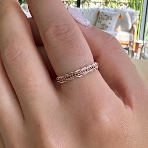 Puede incluir: Un anillo de oro con un diseño trenzado y pequeñas piedras preciosas rosas. El anillo está en el dedo de una persona.