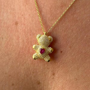 Könnte beinhalten: Goldfarbene Halskette mit einem Teddybär-Anhänger. Der Bär ist mit kleinen, klaren Steinen besetzt und hat ein rotes herzförmiges Zentrum. Die Halskette ist filigran und ebenfalls goldfarben.