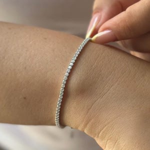 Puede incluir: Una delicada pulsera de plata adornada con numerosas piedras preciosas peque&ntilde;as y brillantes. La pulsera se est&aacute; abrochando en una mu&ntilde;eca, mostrando su elegante dise&ntilde;o y sus intrincados detalles. Las piedras preciosas est&aacute;n muy juntas, creando una l&iacute;nea continua de brillo.
