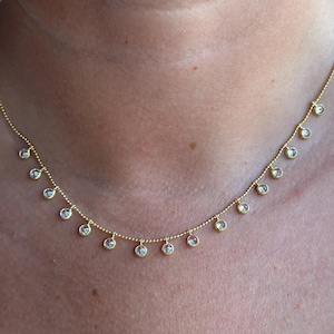 Może przedstawiać: Delikatny złoty naszyjnik z małymi, przezroczystymi kamieniami szlachetnymi równomiernie rozmieszczonymi wzdłuż łańcuszka. Naszyjnik jest noszony na szyi, prezentując swój prosty, ale elegancki design. Kamienie szlachetne wydają się okrągłe i są osadzone w delikatnej złotej oprawie.