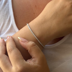 Puede incluir: Una delicada pulsera plateada con numerosas piedras peque&ntilde;as y transparentes se lleva en la mu&ntilde;eca. La pulsera brilla sobre la piel. La persona lleva una camiseta sin mangas blanca.