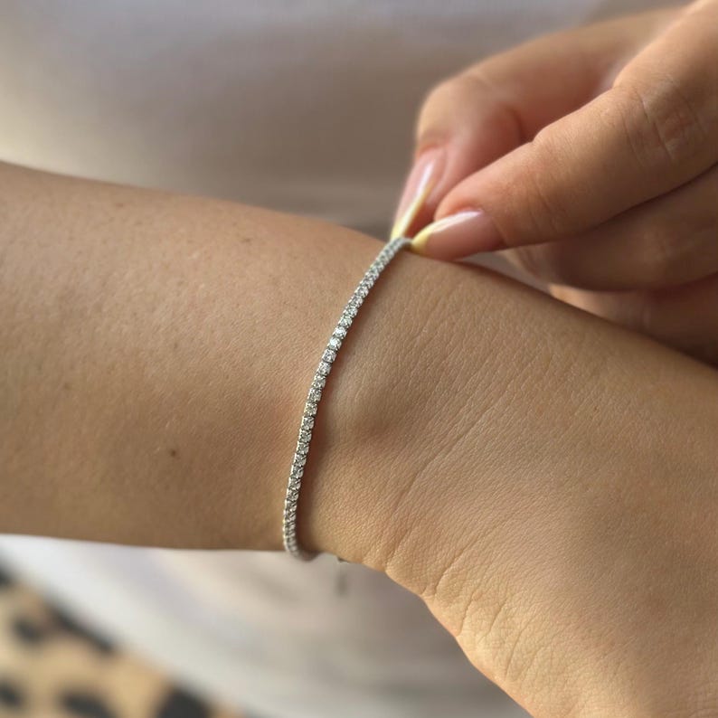 Puede incluir: Una delicada pulsera de plata adornada con numerosas piedras preciosas peque&ntilde;as y brillantes. La pulsera se est&aacute; abrochando en una mu&ntilde;eca, mostrando su elegante dise&ntilde;o y sus intrincados detalles. Las piedras preciosas est&aacute;n muy juntas, creando una l&iacute;nea continua de brillo.