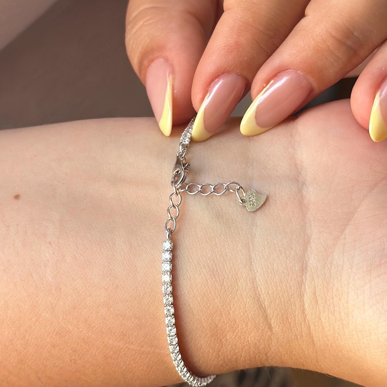 Puede incluir: Una pulsera plateada con una fila de piedras preciosas claras y brillantes. La pulsera se est&aacute; abrochando en una mu&ntilde;eca. El cierre tiene un peque&ntilde;o dije en forma de coraz&oacute;n. El fondo es de color claro y neutro.