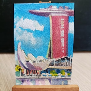 以下が含まれることがあります： シンガポールのマリーナベイ・サンズホテルを描いた水彩画。象徴的な赤いタワーと船の形をした屋根、そして蓮の花の形をした白いアートサイエンス・ミュージアムが、白い雲が浮かぶ明るい青空の下に描かれています。