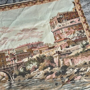 May include: Tapestry featuring a detailed depiction of a European cityscape with a river and bridge in the foreground. The artwork showcases buildings with red-tiled roofs and a large, round fortress. The color palette includes muted greens, browns, and pinks.