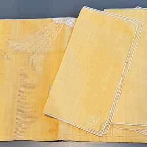 May include: A set of yellow linen table linens. The set includes a rectangular tablecloth with a detailed embroidered design and four matching napkins with white trim. A small red tag is visible on one napkin.