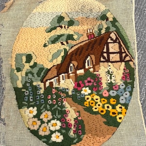 May include: Oval needlepoint of a cottage scene. The cottage has a brown thatched roof, white walls, and a chimney. Colorful flowers line the path leading to the cottage. The background features trees and a light blue sky.