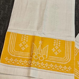 May include: A rectangular off-white linen towel featuring a wide horizontal band of vibrant yellow at the bottom. The yellow section displays a white embroidered trident emblem, geometric floral patterns, and parallel lines, with a hemmed edge.
