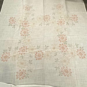 May include: A white linen tablecloth featuring a floral design. The pattern includes peach-colored flowers with green leaves and stems, arranged in a symmetrical layout. The tablecloth is rectangular and appears to be folded.