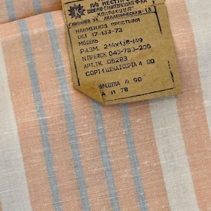 May include: A white fabric featuring vertical stripes in light peach and pale blue. A rectangular tan paper tag is attached, printed with black Cyrillic text including "ММП МССР", "П/О МЕСТПРОМ", "ШВЕЙНО-ГАЛАНТЕРЕЙНАЯ Ф-КА", "КОНФЕКЦИЯ", and dimensions "214x138-149".