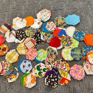 May include: A collection of numerous fabric hexagons in various colors and patterns, including floral, geometric, and solid prints. The pieces are scattered across the surface, showcasing a wide array of textile designs and vibrant hues.