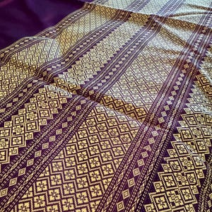 May include: A close-up of a folded textile with a geometric pattern in cream and purple. The fabric has a repeating diamond and square design with vertical stripes. The background is a solid purple fabric.