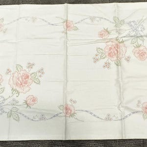 May include: White rectangular fabric with a floral design. Pink roses and green leaves are arranged in a border pattern with light blue ribbons and accents. The fabric is likely a tablecloth or bedding.