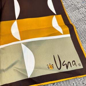 May include: A square scarf featuring a geometric pattern of orange, mustard yellow, and sage green rectangular blocks separated by dark brown borders. The design includes white crescent shapes and a small yellow ladybug icon near the signature Vana.