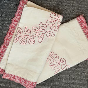 May include: Off-white pillowcases with red embroidered floral designs. The edges are trimmed with a delicate red lace. The pillowcases are folded and rest on a gray surface.