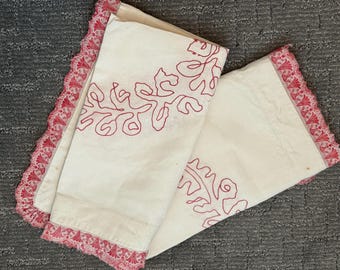 May include: Off-white pillowcases with red embroidered floral designs. The edges are trimmed with a delicate red lace. The pillowcases are folded and rest on a gray surface.