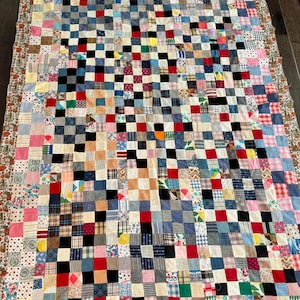 May include: A rectangular patchwork quilt constructed from numerous small, multi-colored fabric squares in various patterns, including solids, plaids, stripes, and floral prints, bordered by a thin strip of brown fabric with a repeating floral motif.