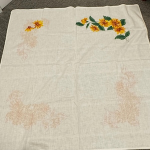 May include: Off-white linen tablecloth with floral embroidery. The corners feature clusters of yellow and orange flowers with green leaves. The other corners have a faded peach-colored floral design. The tablecloth is square.