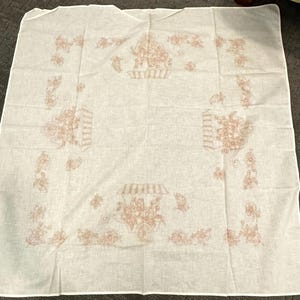 May include: A white tablecloth with a repeating floral design in a muted rose color. The pattern includes floral baskets, floral arrangements, and floral accents. The tablecloth is square and appears to be made of a lightweight fabric.