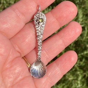 May include: A silver charm in the shape of a spoon with a shell-shaped bowl and ornate detailing. The charm has a green gemstone set in the top.