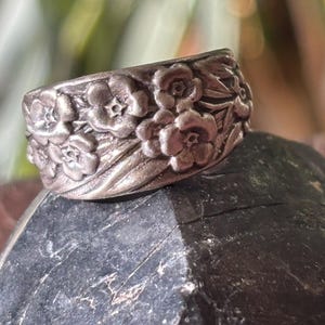 May include: A silver-toned ring with a floral design. The ring features a band covered in detailed, raised flowers. The flowers are clustered together, creating a textured surface. The ring is set against a dark, textured background.