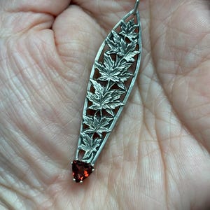 Op de afbeelding: Een zilveren ketting met een delicate hanger met een ontwerp van esdoornbladeren en een rode edelsteen aan de onderkant.