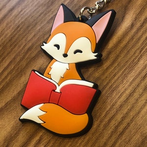 May include: A red and orange fox keychain with black eyes and a white belly. The fox is holding a red book with a white spine. The keychain is made of rubber.
