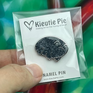 May include: An enamel pin in a clear plastic sleeve. The pin is a dark gray, oval-shaped design with a silver outline, depicting a stylized creature. The packaging includes the text "Kieutie Pie" and "NAMEL PIN".
