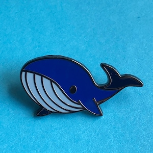 May include: A blue whale enamel pin with a white belly and a black outline. The whale is facing left and has a small, black eye.