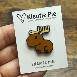 May include: A brown enamel pin featuring a cartoon moose with yellow antlers. The pin is on a white card with the text "Kieutie Pie" and "ENAMEL PIN". The website www.KieutiePie.com is also visible.