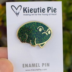 May include: Green glitter enamel pin of a cartoon frog with a sad expression. The pin is gold-toned and has the text "Kieuitie Pie" and "Making Art for the Young at Heart" on the card it is displayed on. The card also says "Enamel Pin" and "www.KieuitiePie.com".
