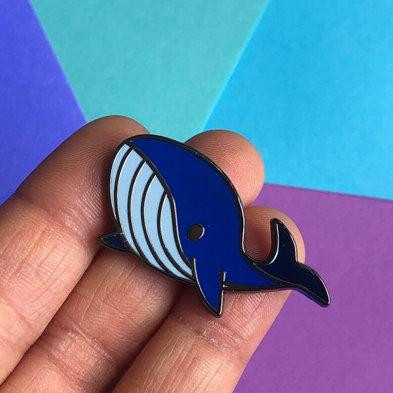 May include: A blue enamel pin shaped like a whale with a white belly and a black eye. The whale is facing the right side of the image.