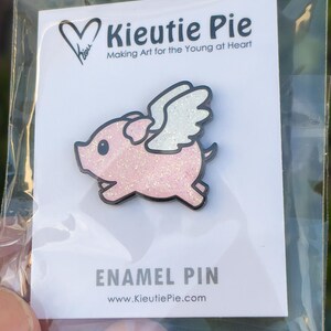 May include: A pink glitter enamel pin of a pig with white wings. The pin says "Kieuitie Pie" and "Making Art for the Young at Heart". It also says "Enamel Pin" and "www.KieuitiePie.com".