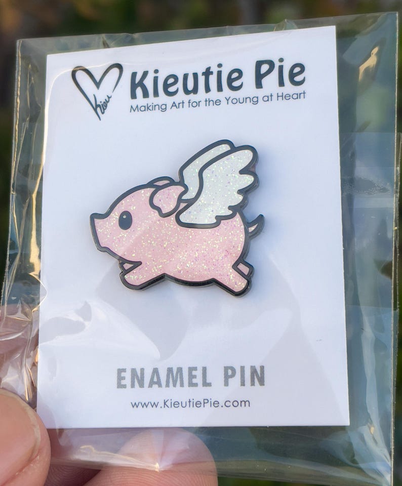 May include: A pink glitter enamel pin of a pig with white wings. The pin says "Kieuitie Pie" and "Making Art for the Young at Heart". It also says "Enamel Pin" and "www.KieuitiePie.com".