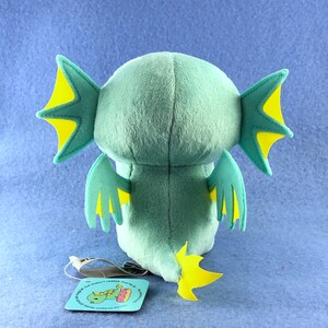 May include: A plush toy of a green and yellow dragon with large wings and a tail. The dragon is facing away from the camera and has a small tag attached to its tail.
