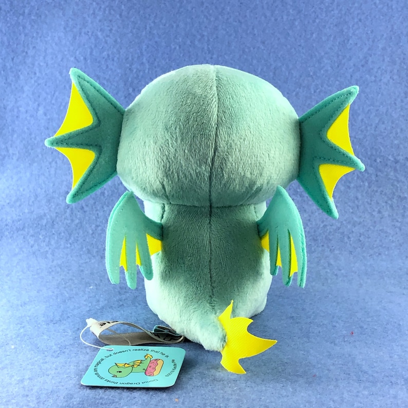 May include: A plush toy of a green and yellow dragon with large wings and a tail. The dragon is facing away from the camera and has a small tag attached to its tail.