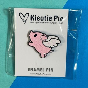 May include: Pink enamel pin featuring a cartoon pig with wings. The pig is wearing a pink glitter finish. The pin says "Kieutie Pie" and "Making Art for the Young at Heart".
