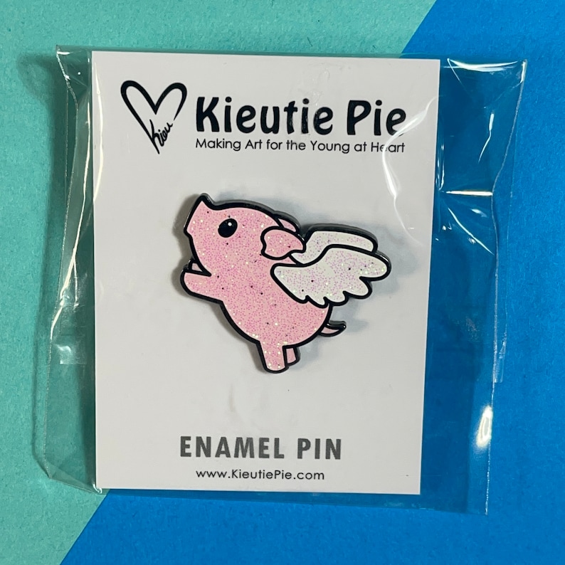 May include: Pink enamel pin featuring a cartoon pig with wings. The pig is wearing a pink glitter finish. The pin says "Kieutie Pie" and "Making Art for the Young at Heart".