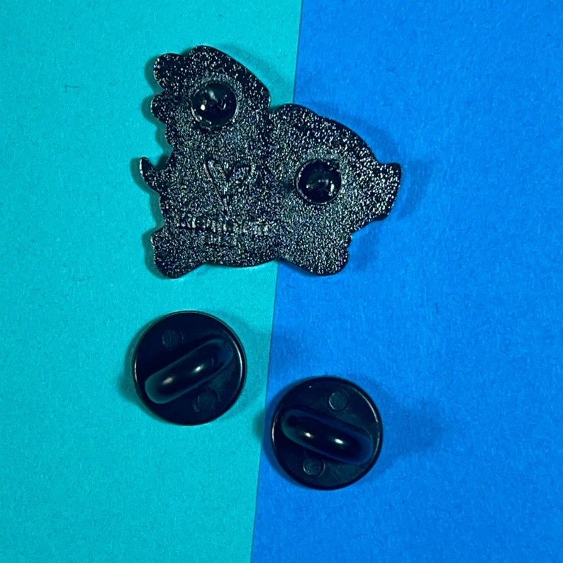 May include: Black enamel pin of a cartoon pig wearing a crown. The pin has two black rubber backings.