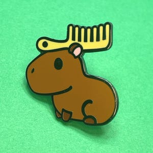 May include: A brown capybara enamel pin with a yellow comb-shaped accessory on its head. The pin has a black outline and pink accents. The capybara is smiling, and the background is a solid green.