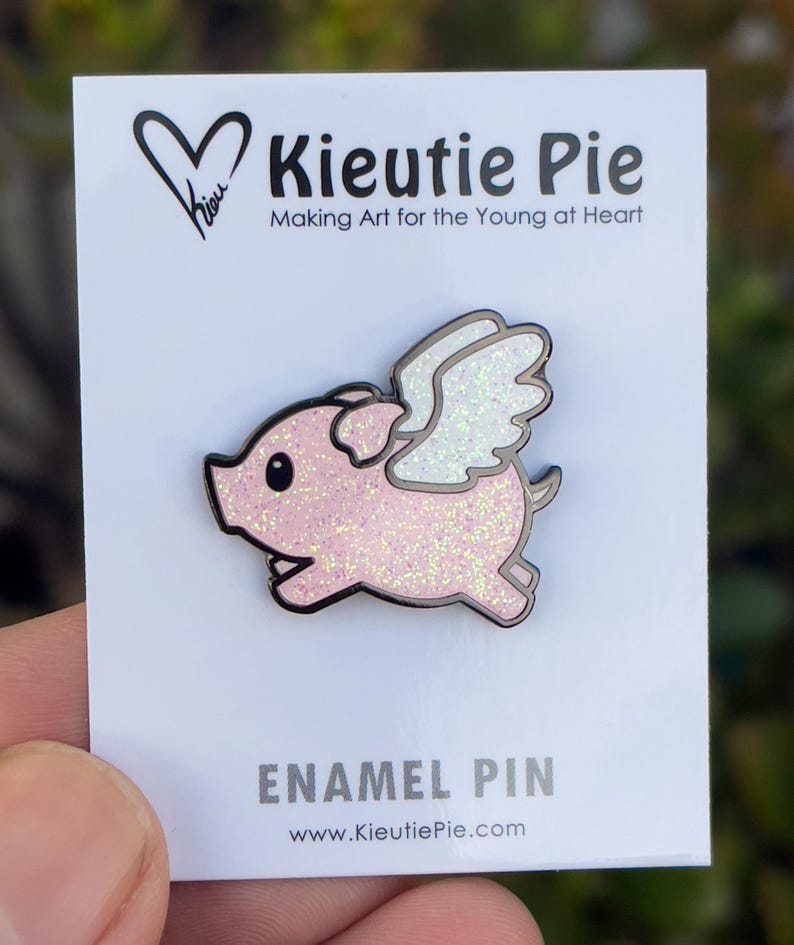 May include: Pink enamel pin of a flying pig with glitter. The pig has white wings and black outlines. The pin is on a white card with the text "Kieutie Pie Making Art for the Young at Heart" and "Enamel Pin www.KieutiePie.com".