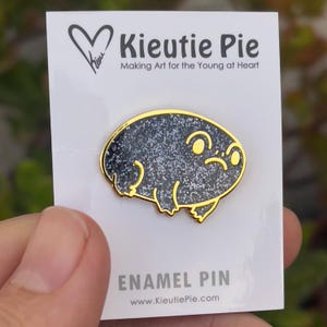 May include: A black and gold enamel pin featuring a cartoon frog with a glittery finish. The pin is on a white card with the text "Kieutie Pie Making Art for the Young at Heart" and "ENAMEL PIN www.KieutiePie.com".