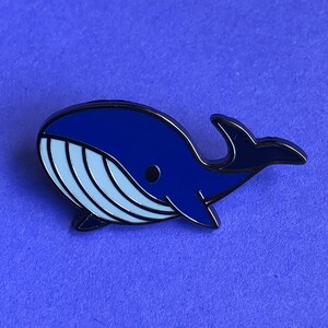 May include: A blue enamel pin shaped like a whale with a white belly and black outline.