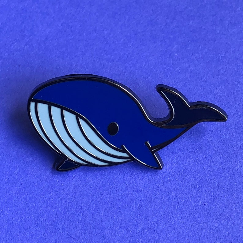 May include: A blue enamel pin shaped like a whale with a white belly and black outline.