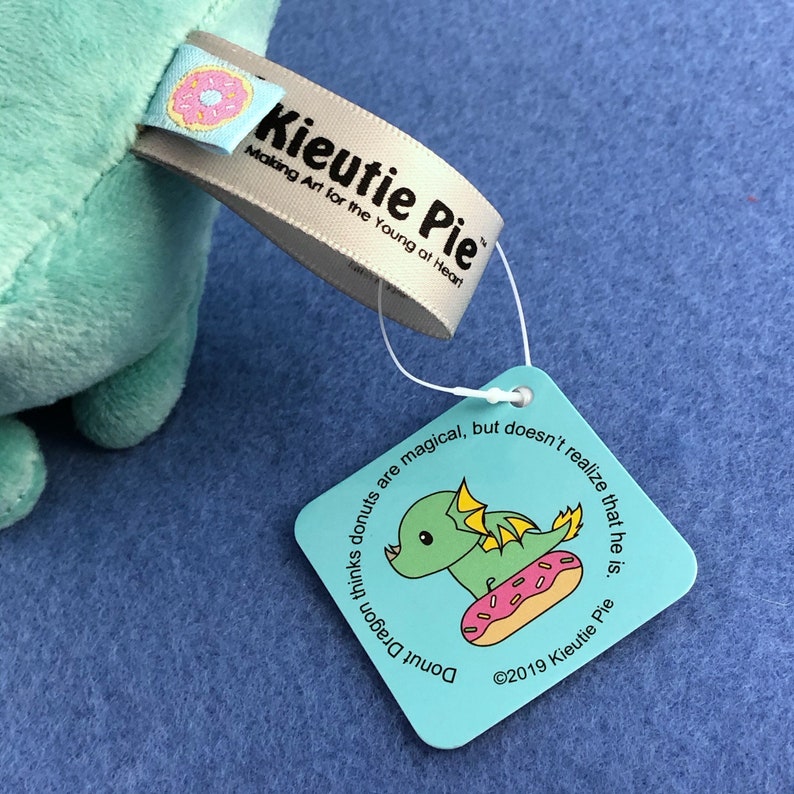 May include: A light blue plush toy shaped like a dragon sitting on a donut. The toy has a tag that reads "Kieutie Pie Making Art for the Young at Heart" and a smaller tag that says "Donut Dragon thinks donuts are magical, but doesn't realize that he is. &copy;2019 Kieutie Pie"