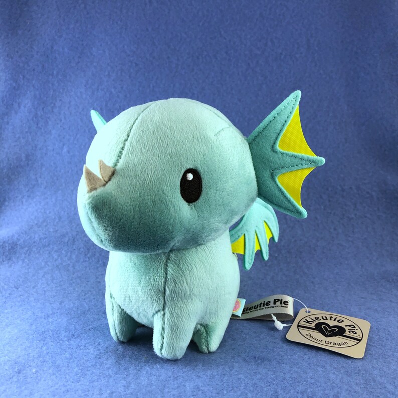 May include: A teal blue plush toy dragon with yellow wings and black eyes. The dragon is standing on a blue surface and has a tag that says "Kutie Pie Donut Dragon".