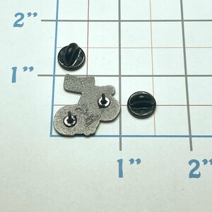 May include: A silver-colored enamel pin in the shape of a stocking, with two black pin backs. The pin is approximately 1 inch in length. The back of the pin has the text "&copy;2023 Katie Me" and a heart symbol.
