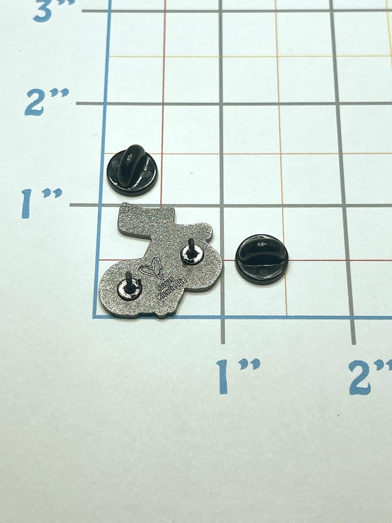 May include: A silver-colored enamel pin in the shape of a stocking, with two black pin backs. The pin is approximately 1 inch in length. The back of the pin has the text "&copy;2023 Katie Me" and a heart symbol.